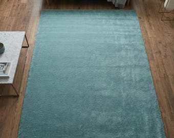 May include: A rectangular area rug in a solid, muted teal blue color with a soft, plush, high-pile texture. The rug is positioned centrally on a wooden floor, covering a large portion of the visible space in front of a sofa and a marble table.