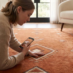 Hand tufted wool rug in rich terracotta featuring a southwestern tribal geometric pattern. Bold diamond medallion centre with concentric borders, ivory and charcoal accents, aztec-inspired motifs. Plush medium pile, artisan crafted, placed on warm oak hardwood floor. Perfect for boho living room, rustic bedroom or as a unique housewarming gift. Custom sizes available.