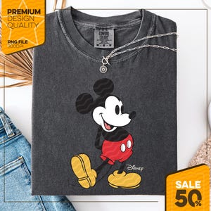 May include: Dark gray Comfort Colors t-shirt featuring a smiling Mickey Mouse graphic. Mickey is wearing red shorts and yellow shoes. A silver necklace with a pendant is on the shirt. The shirt has the Disney logo and the Comfort Colors label.