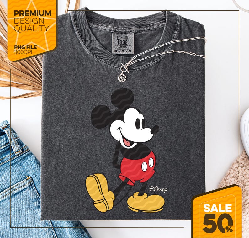 May include: Dark gray Comfort Colors t-shirt featuring a smiling Mickey Mouse graphic. Mickey is wearing red shorts and yellow shoes. A silver necklace with a pendant is on the shirt. The shirt has the Disney logo and the Comfort Colors label.