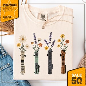 May include: Cream-colored t-shirt featuring a floral design. The graphic depicts four stylized lightsabers, each holding a different type of flower. The shirt has the words "Comfort Colors" on the tag. A gold necklace is draped across the shirt. The image also includes the text "Premium Design Quality" and "Sale 50%"