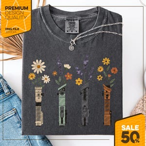 May include: Dark gray t-shirt with a floral design. The design features four stylized lightsabers with flowers blooming from the top. The shirt has the text "Comfort Colors" and "Premium Design Quality". A silver necklace is also visible.