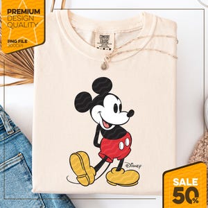 May include: Cream-colored t-shirt featuring a graphic of Mickey Mouse in his classic pose. Mickey is depicted in black, red, yellow, and white. The shirt has the Disney logo and the words "Comfort Colors" on the tag.