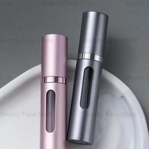 K&ouml;nnte beinhalten: Zwei metallische Parf&uuml;mzerst&auml;uber, einer in zartem Rosa und der andere in Silbergrau, werden auf einem wei&szlig;en, marmorierten Tablett pr&auml;sentiert. Jeder Zerst&auml;uber hat ein rechteckiges Fenster und eine passende Kappe.