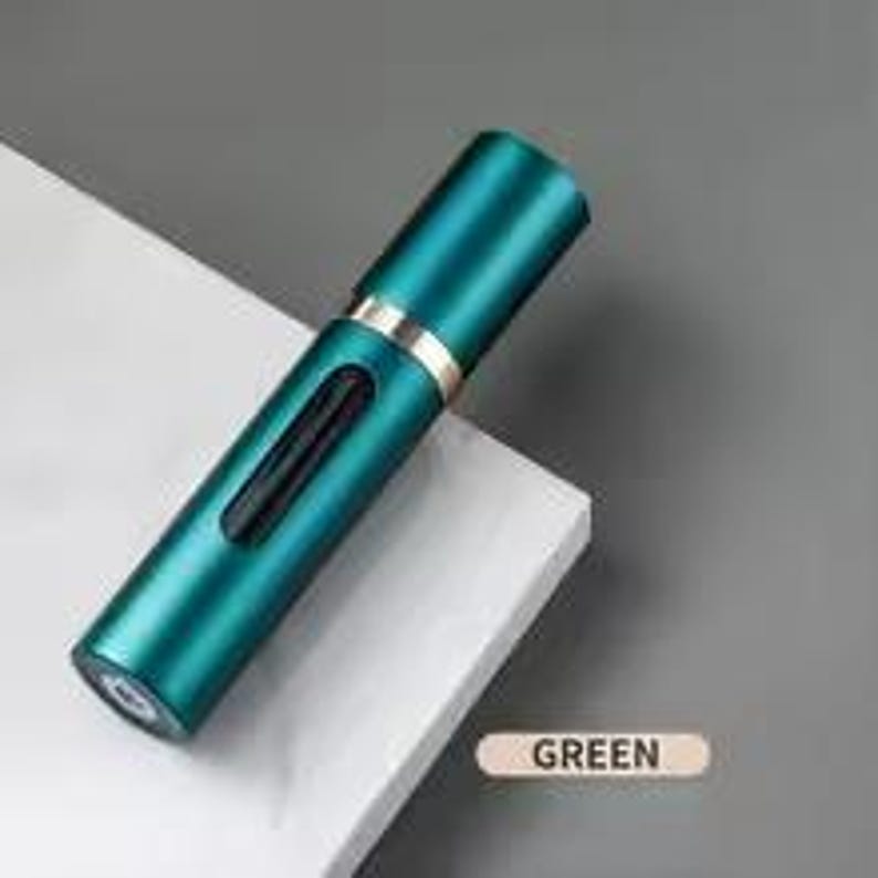 K&ouml;nnte beinhalten: Ein zylindrischer, teal-farbener Parf&uuml;mzerst&auml;uber mit einem goldfarbenen Band. Der Zerst&auml;uber hat ein kleines, rechteckiges Fenster und eine Spr&uuml;hd&uuml;se. Das Wort "GREEN" ist auf einem kleinen, rechteckigen Etikett aufgedruckt.