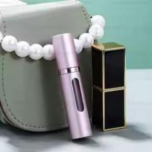 K&ouml;nnte beinhalten: Ein hellrosa Parf&uuml;mzerst&auml;uber mit silbernem Band und rechteckigem Fenster, neben einem schwarzen und goldenen Lippenstiftetui. Eine graue Handtasche und eine wei&szlig;e Perlenkette sind im Hintergrund.