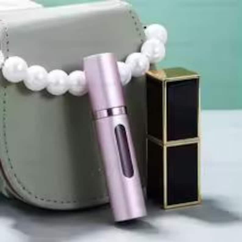 K&ouml;nnte beinhalten: Ein hellrosa Parf&uuml;mzerst&auml;uber mit silbernem Band und rechteckigem Fenster, neben einem schwarzen und goldenen Lippenstiftetui. Eine graue Handtasche und eine wei&szlig;e Perlenkette sind im Hintergrund.