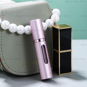 K&ouml;nnte beinhalten: Ein rosa Parf&uuml;mzerst&auml;uber mit einem transparenten Fenster, ein schwarzer und goldener Lippenstift und eine gr&uuml;ne Handtasche mit Perlenriemen. Der Zerst&auml;uber ist zylindrisch und hat ein metallisches Finish. Der Lippenstift ist rechteckig. Auf der Handtasche steht das Wort "Secret".
