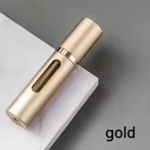 K&ouml;nnte beinhalten: Ein goldfarbener, zylindrischer Parf&uuml;mzerst&auml;uber mit einem kleinen, rechteckigen Fenster. Der Zerst&auml;uber hat eine Kappe und eine Spr&uuml;hd&uuml;se. Das Wort "gold" ist unten rechts aufgedruckt.