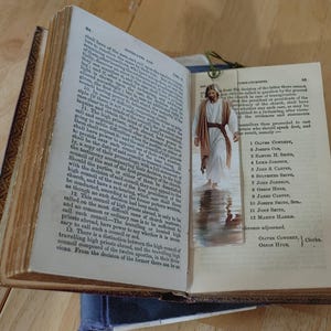 May include: An open antique book with aged, yellowed pages. A bookmark featuring an image of Jesus walking on water is inserted. The book's cover has a dark, ornate design. The pages display printed text.