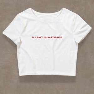 May include: White crop top t-shirt with short sleeves. The red text on the shirt reads "IT'S THE TEQUILA TALKING". The shirt has a round neckline and is made of a soft, lightweight fabric.
