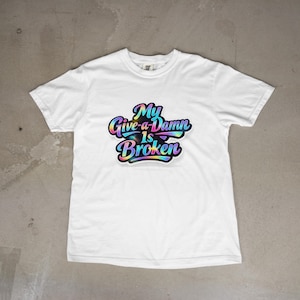 Op de afbeelding: Wit T-shirt met de tekst "My Give-a-Damn Is Broken" in een kleurrijk, gestileerd lettertype. De tekst heeft een regenbooggradiënteffect. Het T-shirt is gemaakt van een zacht materiaal en is geschikt voor casual wear.