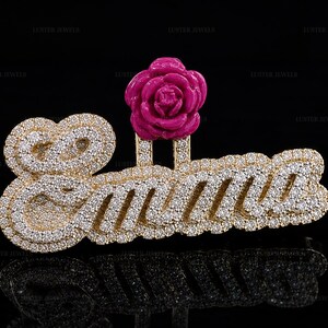 May include: A gold-toned pendant featuring the name Emma in cursive script, encrusted with numerous small clear stones. A bright pink rose-shaped accent is attached to the top of the letter m, creating a decorative floral detail on the jewelry.