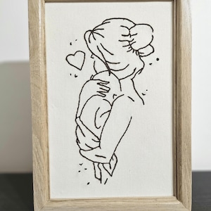 Puede incluir: Una obra de arte bordada enmarcada que presenta un dibujo lineal minimalista de una persona abrazando a un niño. La obra está realizada en hilo negro sobre lienzo blanco, con un marco de madera. También se borda un corazón y pequeños puntos.