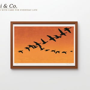 May include: A framed art print featuring a flock of black geese silhouettes flying in a V-formation against a vibrant orange gradient background. The artwork is enclosed in a simple brown wooden frame, presenting a clear contrast between the dark birds and the warm, sunset-coloured sky depicted in the print.
