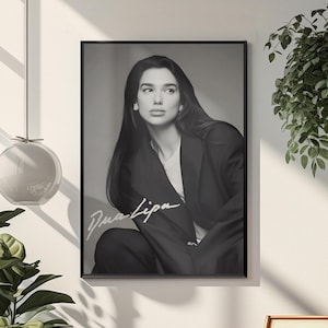 Può includere: Ritratto in bianco e nero incorniciato di Dua Lipa. L'immagine la mostra con un abito scuro e lunghi capelli scuri. La firma dell'artista è visibile nell'angolo in basso a sinistra. Il ritratto è appeso a una parete bianca.