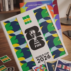 Puede incluir: Un álbum de cromos de la Copa Mundial de la FIFA 2026 con cubierta blanca, un gran logotipo 26 en negro, el trofeo de la FIFA y patrones geométricos coloridos. Incluye sobres de cromos y pegatinas con el texto WE ARE BRA y World Cup 2026.