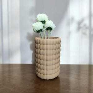 Pode incluir: Um vaso bege texturizado com um design arredondado em forma de bolha, segurando três flores brancas com caules verdes. O vaso está sobre uma superfície de madeira. O vaso tem cerca de 15 cm de altura.