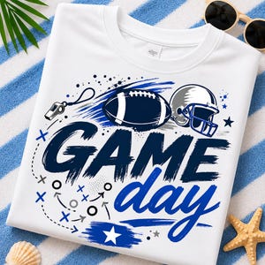 Puede incluir: Una camiseta blanca con un estampado gráfico que incluye el texto "GAME day" en letras azul oscuro y azul con efecto de pincelada. El diseño muestra un balón de fútbol americano azul oscuro, un casco plateado, un silbato y diagramas de juego.