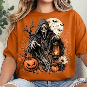 May include: An orange t-shirt featuring a graphic of a hooded figure with a white mask holding a lit lantern. The scene includes a full moon with bats, bare tree branches, autumn leaves, and two jack-o'-lanterns at the base of the figure.