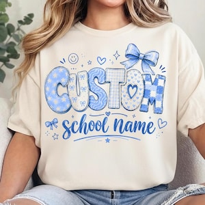 May include: A cream-colored t-shirt featuring a blue graphic design with the text CUSTOM school name. The design includes various blue patterns like stars, plaid, and bows, along with small heart, star, and smiley face icons and a large blue bow.