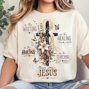 Puede incluir: Una camiseta de color crema con un gráfico de una cruz negra cubierta con tela, rodeada de arreglos florales y mariposas. El texto dice: in the WAITING, in the HEALING, in the HURTING, in the SEARCHING, there was JESUS.