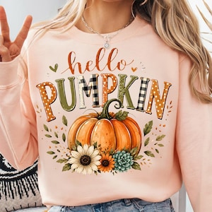 Puede incluir: Una sudadera de cuello redondo en color melocotón claro con el texto hello PUMPKIN en una mezcla de patrones naranjas, verdes y cuadros blancos y negros. Debajo del texto hay una calabaza naranja grande rodeada de girasoles y hojas.