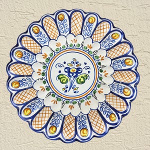 May include: A round ceramic plate with a scalloped edge, featuring a central blue and green floral motif surrounded by concentric rings. The border displays a repeating pattern of orange cross-hatched sections and yellow circular accents.