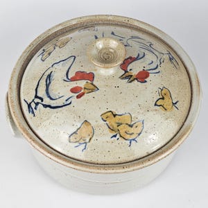 May include: A round ceramic casserole dish with a lid, featuring a speckled beige glaze. The lid is decorated with hand-painted illustrations of a hen with a red comb and three small yellow chicks, finished with a central knob handle.