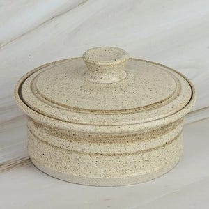 Puede incluir: Un recipiente redondo de cerámica color crema con tapa, que presenta una textura moteada en toda su superficie. El objeto cuenta con un borde ligeramente elevado en la tapa, un pomo central como asa y bandas horizontales sutiles.