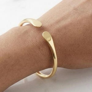 May include: A polished gold-tone cuff bracelet featuring a smooth, open-ended design with rounded, teardrop-shaped terminals. The metal band has a reflective surface and a consistent thickness, curving around the wrist in a simple, continuous loop.