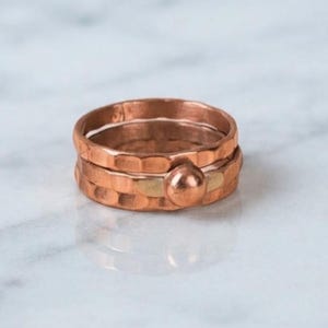 May include: A set of two stacked copper rings featuring a hammered, textured finish. The top ring is a simple band, while the bottom ring includes a small, smooth, round copper bead accent integrated into the hammered metal surface.