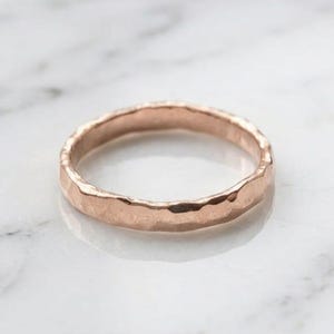 Pode incluir: Um anel de ouro rosa com um acabamento de superfície martelado e texturizado. A banda tem uma largura consistente e um perfil arredondado, exibindo um brilho metálico que reflete a luz através da sua superfície exterior irregular e amolgada.