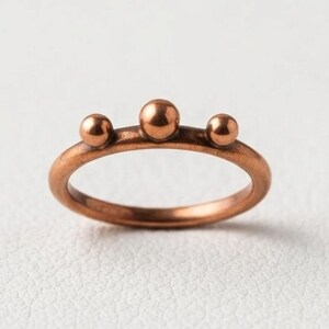 Op de afbeelding: Een koperkleurige metalen ring met een gladde, dunne band, versierd met drie kleine, bolvormige kralen van verschillende groottes die in een rij aan de bovenkant zijn geplaatst. De middelste kraal is iets groter dan de twee zijdelingse.