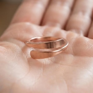 Op de afbeelding: Een koperkleurige bypass-ring met een gladde, gepolijste afwerking. De ring is aan de voorzijde open, met twee licht taps toelopende uiteinden die elkaar overlappen. Het metaal heeft een warme, metallic glans en een eenvoudig, strak ontwerp.