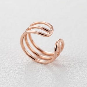 Peut inclure: Une boucle d'oreille ear cuff en or rose au design triple anneau avec une finition lisse et polie. La structure circulaire ouverte se compose de trois fils métalliques parallèles et interconnectés qui se courbent en une forme continue.