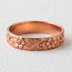 Può includere: Un anello a fascia in oro rosa caratterizzato da un motivo inciso continuo di piccoli fiori a cinque petali e volute vegetali. Il metallo presenta una calda tonalità rosata metallica e una finitura lucida con dettagli floreali complessi.