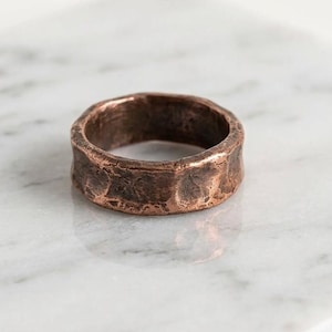 May include: A band ring crafted from copper with a dark, oxidized finish. The surface of the ring features a heavily textured, hammered appearance with irregular indentations and a rugged, uneven edge profile that encircles the entire band.