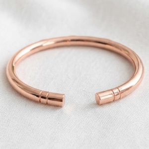 May include: A rose gold-toned cuff bracelet with a smooth, polished finish. The circular band features an open design with two distinct, parallel etched lines encircling each of the rounded, flat-faced ends of the metal cuff.