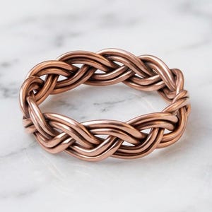 May include: A copper-colored ring featuring a continuous braided design. The band is constructed from multiple strands of metal woven together to create a textured, circular pattern with a warm, metallic finish and a consistent, uniform thickness.