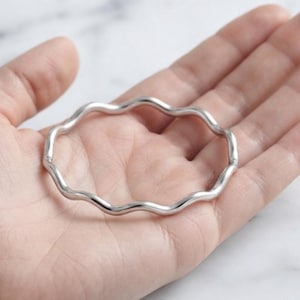 Puede incluir: Un brazalete rígido de tono plateado que presenta un diseño continuo de ondas. El metal tiene un acabado liso y pulido, con una forma circular y un grosor constante a lo largo de toda la circunferencia de la pieza de joyería.