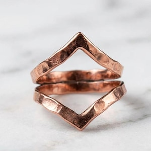 May include: A copper ring featuring a V-shaped chevron design with a hammered, textured finish. The band has an open, angular silhouette that creates a pointed peak at the top, showcasing the metallic reddish-brown color and reflective surface.