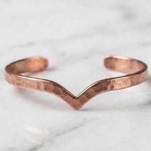 May include: A copper cuff bracelet featuring a V-shaped centre point and a textured, hammered finish. The open-ended band has a metallic reddish-brown hue and a consistent width throughout its design, creating a simple and clean aesthetic.