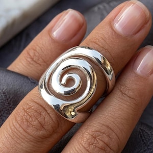 May include: A polished silver ring featuring a prominent openwork spiral design. The band is smooth and reflective, showcasing a continuous, swirling metal pattern that wraps around the finger, creating a bold and eye-catching statement piece.
