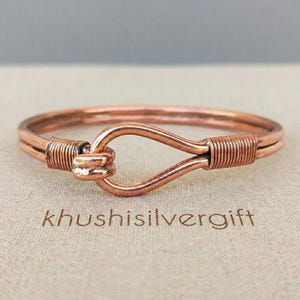 Puede incluir: Una pulsera de alambre color cobre con un cierre de estilo gancho. El diseño consta de dos alambres paralelos asegurados por bobinas de alambre apretadas a ambos lados del cierre central en forma de bucle. El texto khushisilvergift es visible.