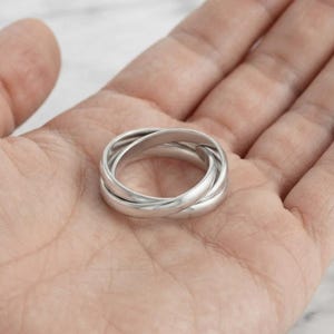 May include: A silver-coloured ring composed of three interlocking bands that overlap to form a single circular structure. The metal surface is smooth and polished, reflecting light across the rounded edges of the connected bands.
