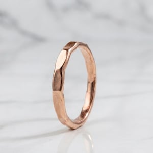 May include: A rose gold ring with a hammered, faceted texture. The band features a series of flat, reflective surfaces around its circumference, creating a geometric appearance with a warm, metallic finish and a consistent, rounded inner profile.