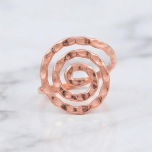 Puede incluir: Un anillo de oro rosa con un diseño de espiral prominente. La superficie metálica tiene un acabado martillado y texturizado que capta la luz, creando una serie de hendiduras a lo largo de la banda circular enrollada hacia el centro.