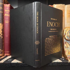 Puede incluir: Un libro de tapa dura negro titulado "The Book of Enoch" con letras doradas. El lomo dice "The Book of Enoch" y "Secret Doctrine Reference Series". El libro se exhibe en una estanter&iacute;a con otros libros.