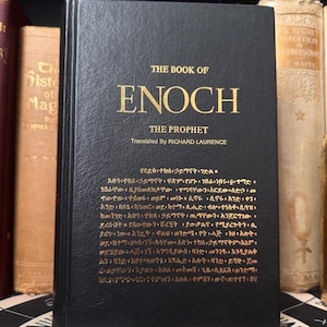 Puede incluir: Un libro de tapa dura negro titulado "The Book of Enoch" con letras doradas. El texto incluye "The Prophet" y "Translated By Richard Laurence." Otros libros son visibles en el fondo.